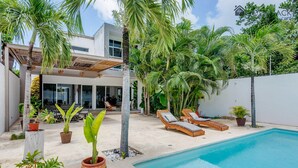 Een moderne woning met een zwembad, lounge stoelen en tropische planten.