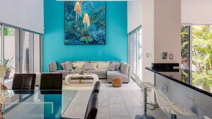Een moderne woonkamer met een blauwgroene accentmuur, een groot abstract schilderij, een grijze bank, een glazen salontafel en een zichtbare keukenruimte door een glazen scheidingswand.