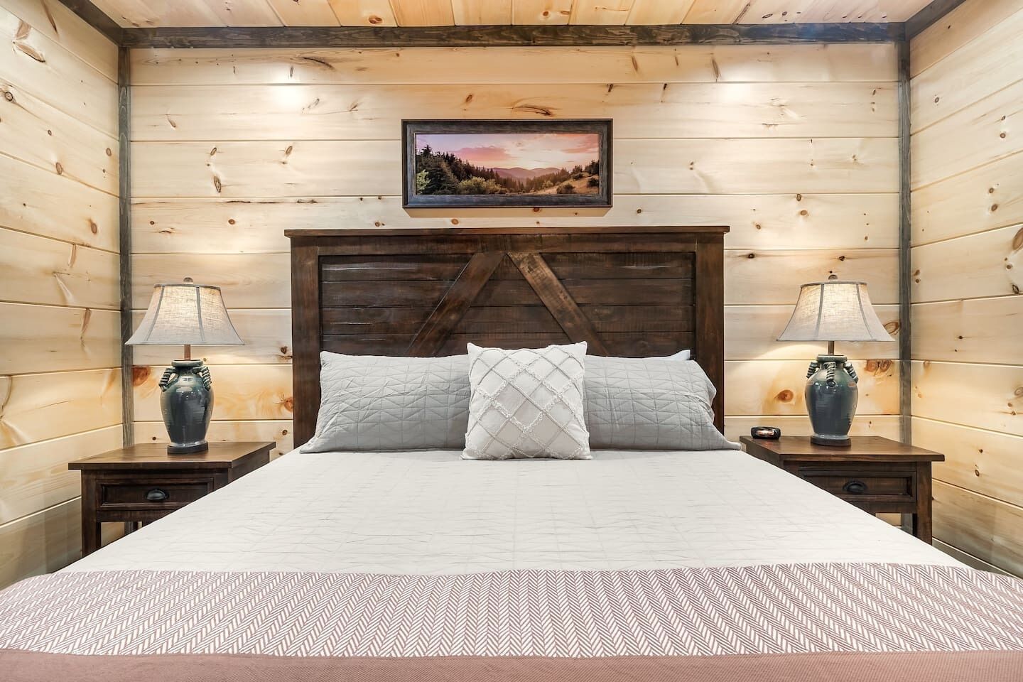 A bedroom with a wooden headboard, a bed with two pillows, two bedside tables with lamps, and a framed picture on the wall.