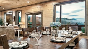 A dining area with a long wooden table set for a meal, large windows with a mountain view, and a cozy seating area with a sofa and chairs. - Private Indoor Heated Pool, Mtn Views, Hot Tub, Fire Pit, Location, Fast Wifi (Sevierville)