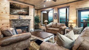 A cozy living room with a stone fireplace, leather sofas, a coffee table, and large windows offering a view of the outdoors. - Private Indoor Heated Pool, Mtn Views, Hot Tub, Fire Pit, Location, Fast Wifi (Sevierville)