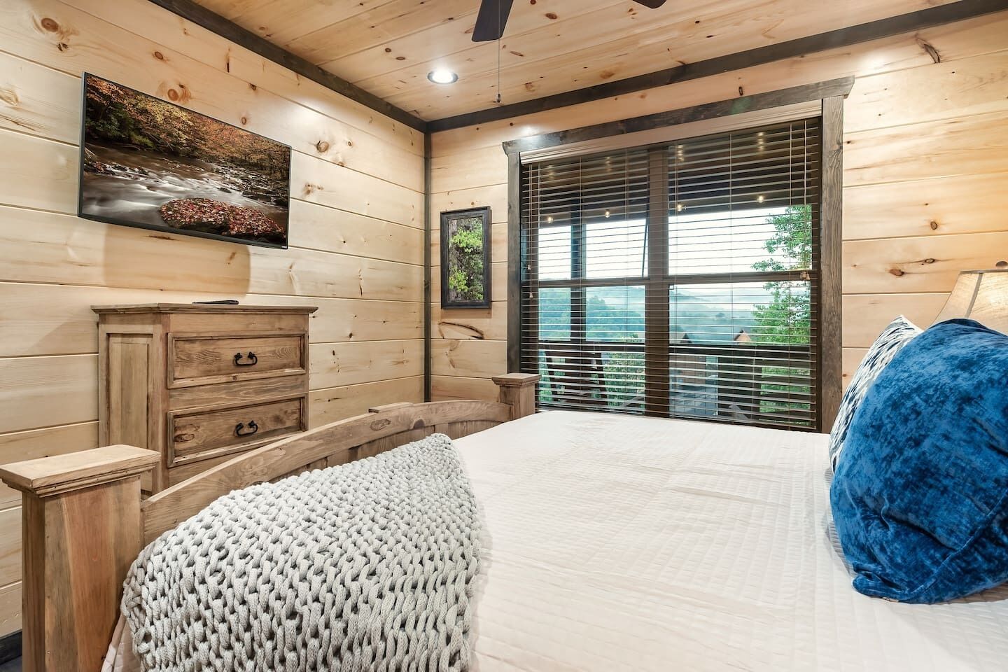 Ein Zimmer mit Holzwänden, einem Bett, einer Kommode, einem Fernseher und einem großen Fenster mit Jalousien.