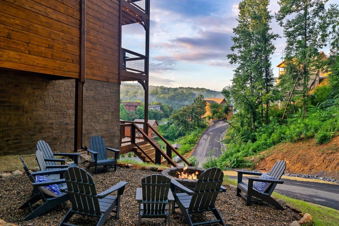 A patio with Adirondack chairs, a fire pit, and a view of a winding road and forested hills.