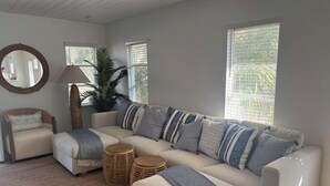 Una sala de estar con un sofá seccional color beige, almohadones a rayas, una mesa de centro redonda de madera, un tapete tejido para el suelo, un espejo redondo y una lámpara de pie alta.