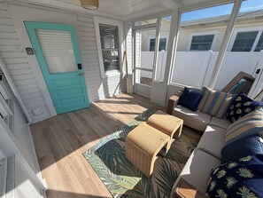 A sunlit room with a teal door, a wooden floor, a sofa with patterned cushions, and a woven basket.