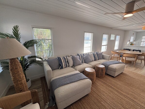A living room with a sectional sofa, a dining table with chairs, a lamp, and a large window with blinds.