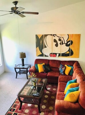 Ein Wohnzimmer mit einem roten Sofa, einem Glastisch, einem Deckenventilator und einem großen abstrakten Gemälde an der Wand.