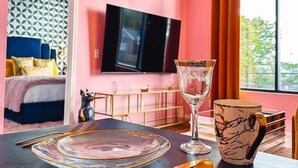 A modern bedroom with a pink wall, a bed with blue bedding, a TV mounted on the wall, and a dining area with a table set for two.