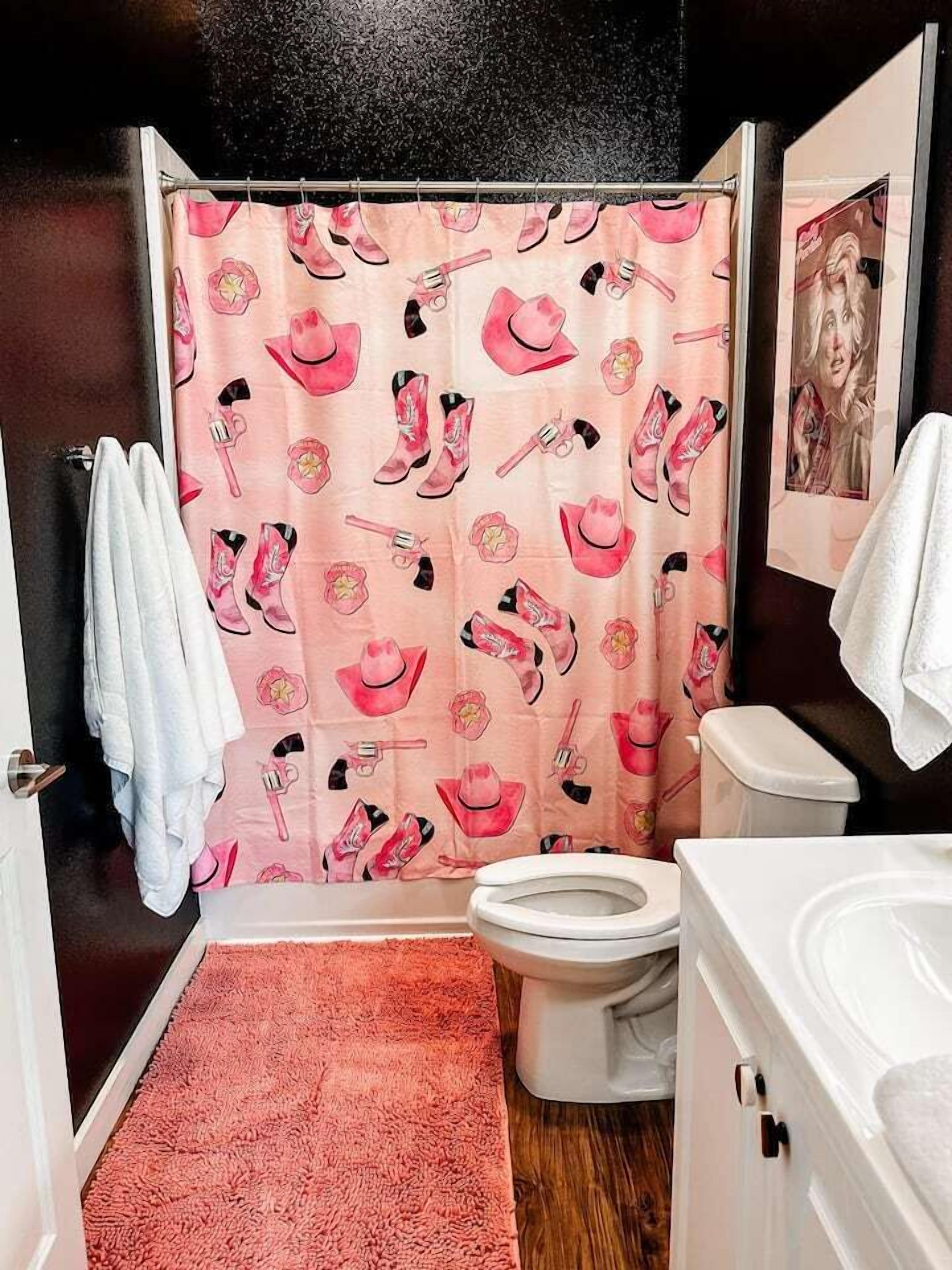 Un baño con cortina de ducha rosa decorada con botas y sombreros de vaquero, un inodoro blanco y un lavamanos blanco.
