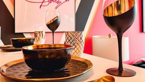 Un coin repas avec une table dressée pour deux personnes, agrémenté d’un bol noir, d’un verre à vin et de baguettes. Une œuvre d’art encadrée est accrochée au mur, et un luminaire moderne est visible.