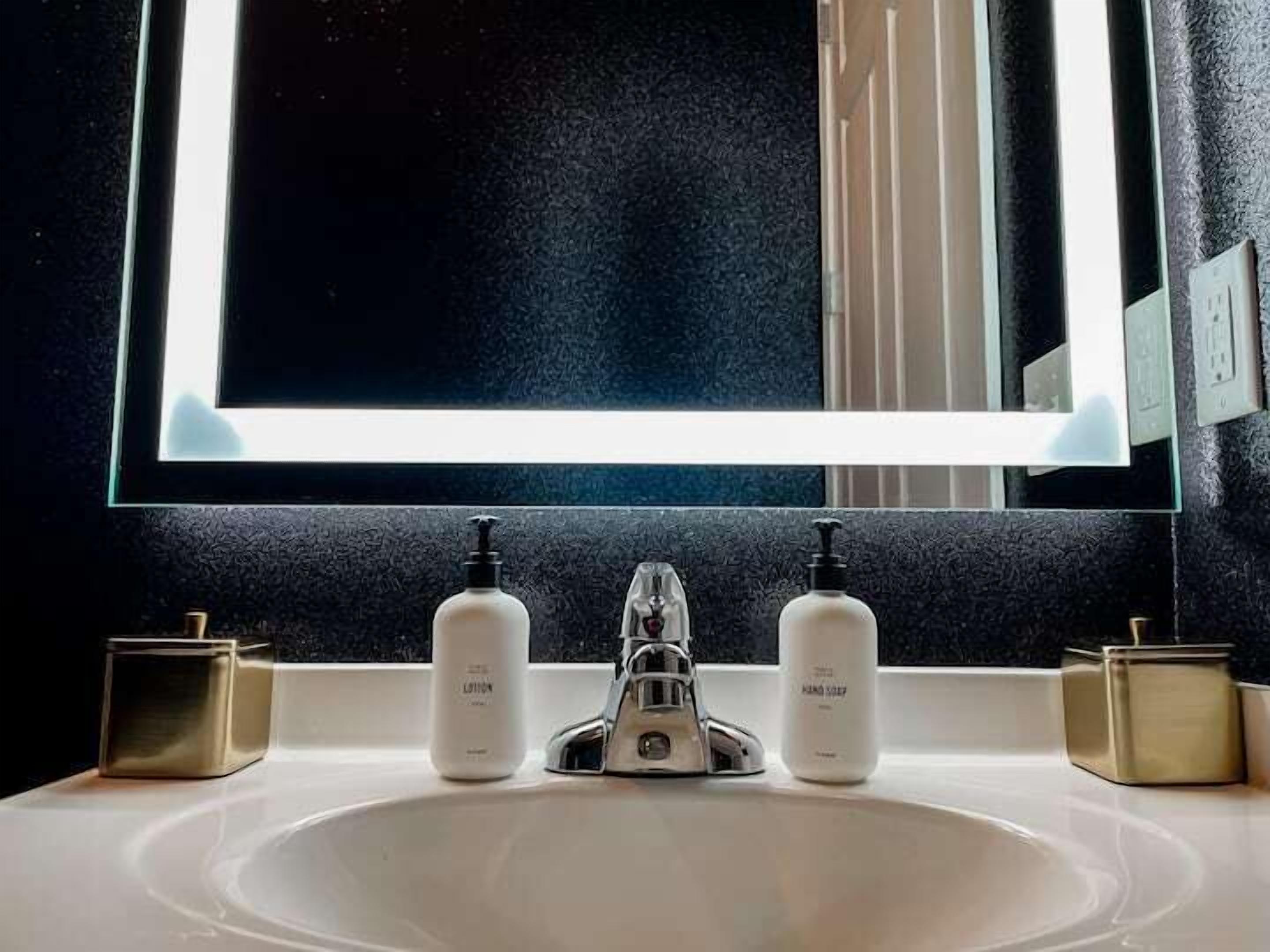 Un baño con lavamanos blanco, dos dispensadores de jabón, un grifo y un espejo con iluminación.