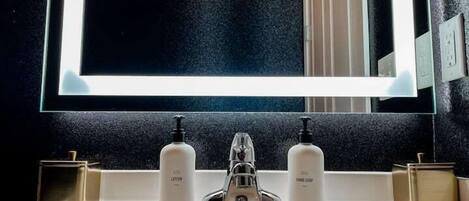 Un baño con lavamanos blanco, dos dispensadores de jabón, un grifo y un espejo con iluminación.