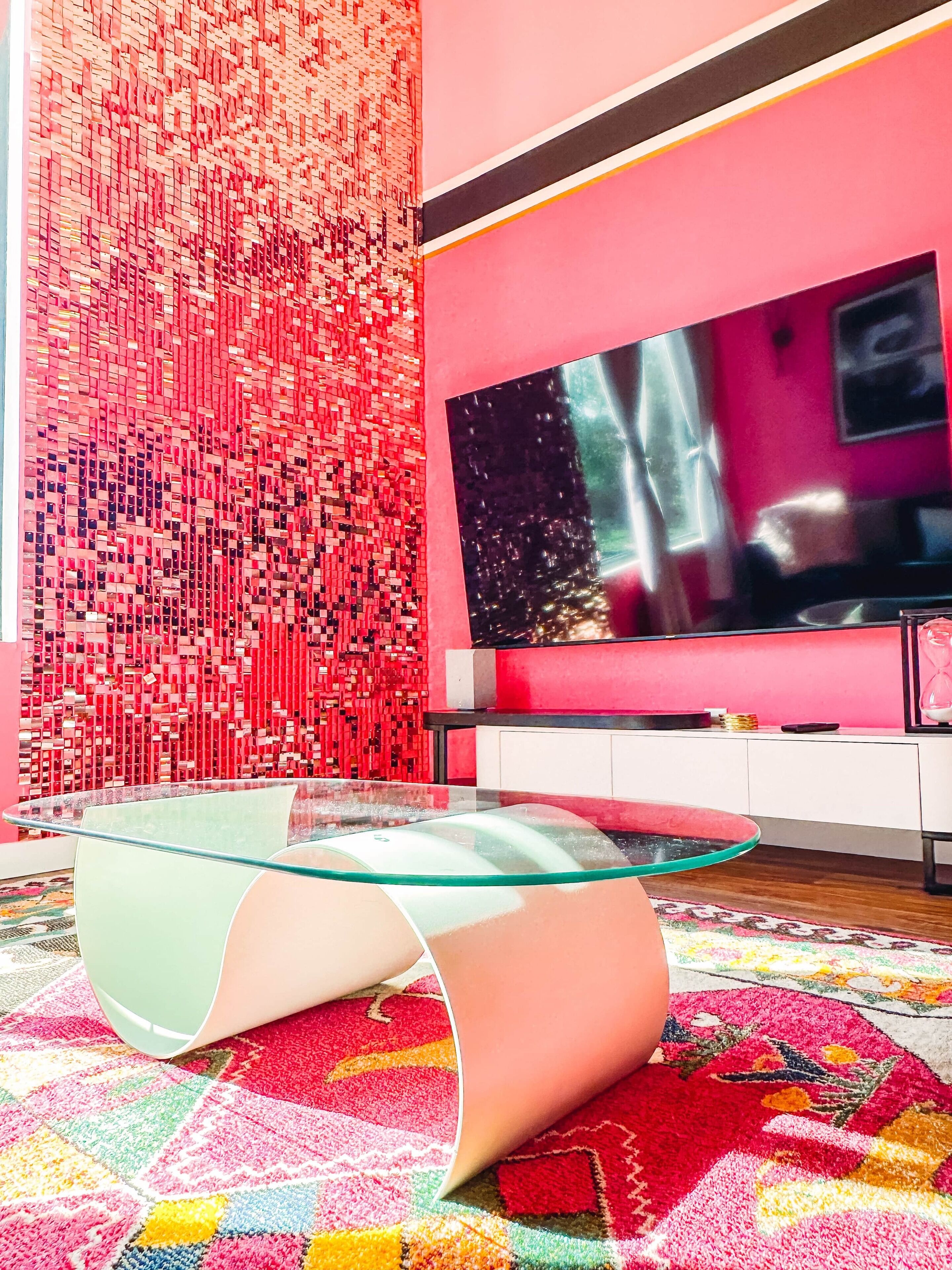 A modern living room with a glass coffee table, a flat-screen TV, and a vibrant red wall with a mosaic pattern.