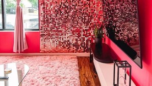 Un salon moderne aux couleurs rose et rouge, avec une grande fenêtre, un tapis rose et moelleux, une table basse en verre, un banc blanc et un panneau décoratif mural.