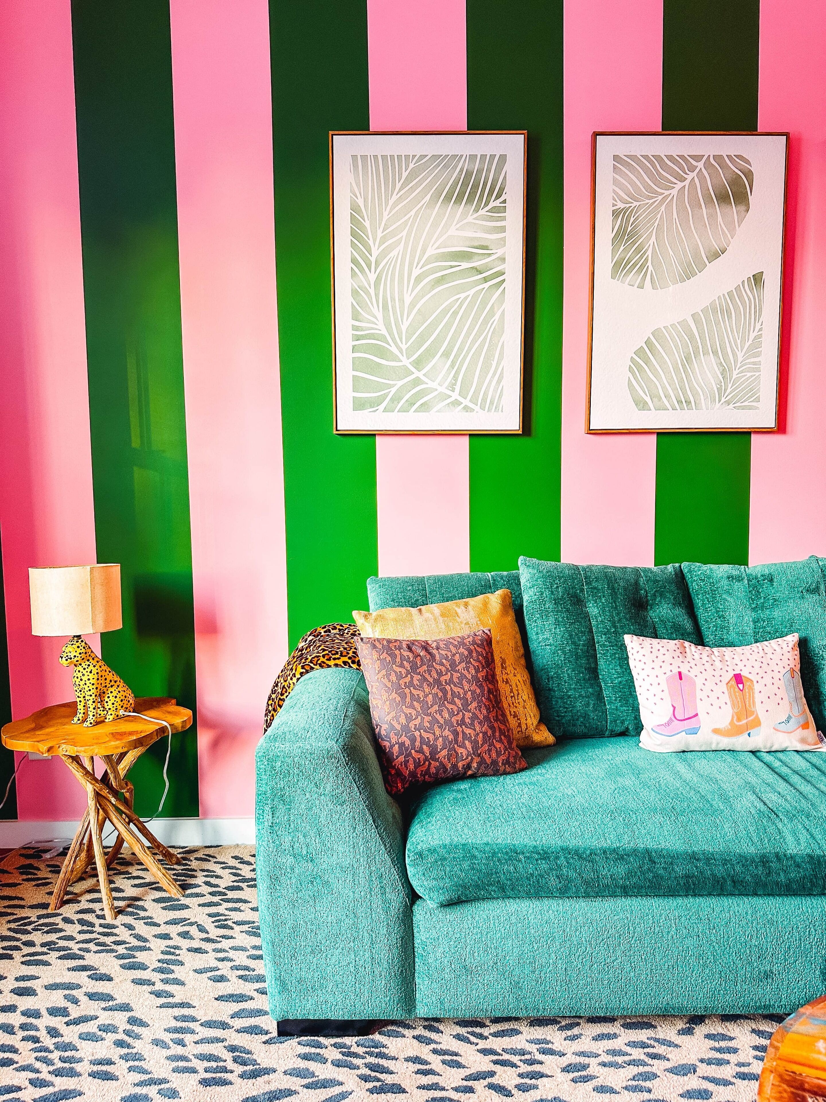 A teal sofa with patterned cushions, a leopard print pillow, and a leopard print lamp on a wooden table. Two framed leaf artworks are on a green and pink striped wall.