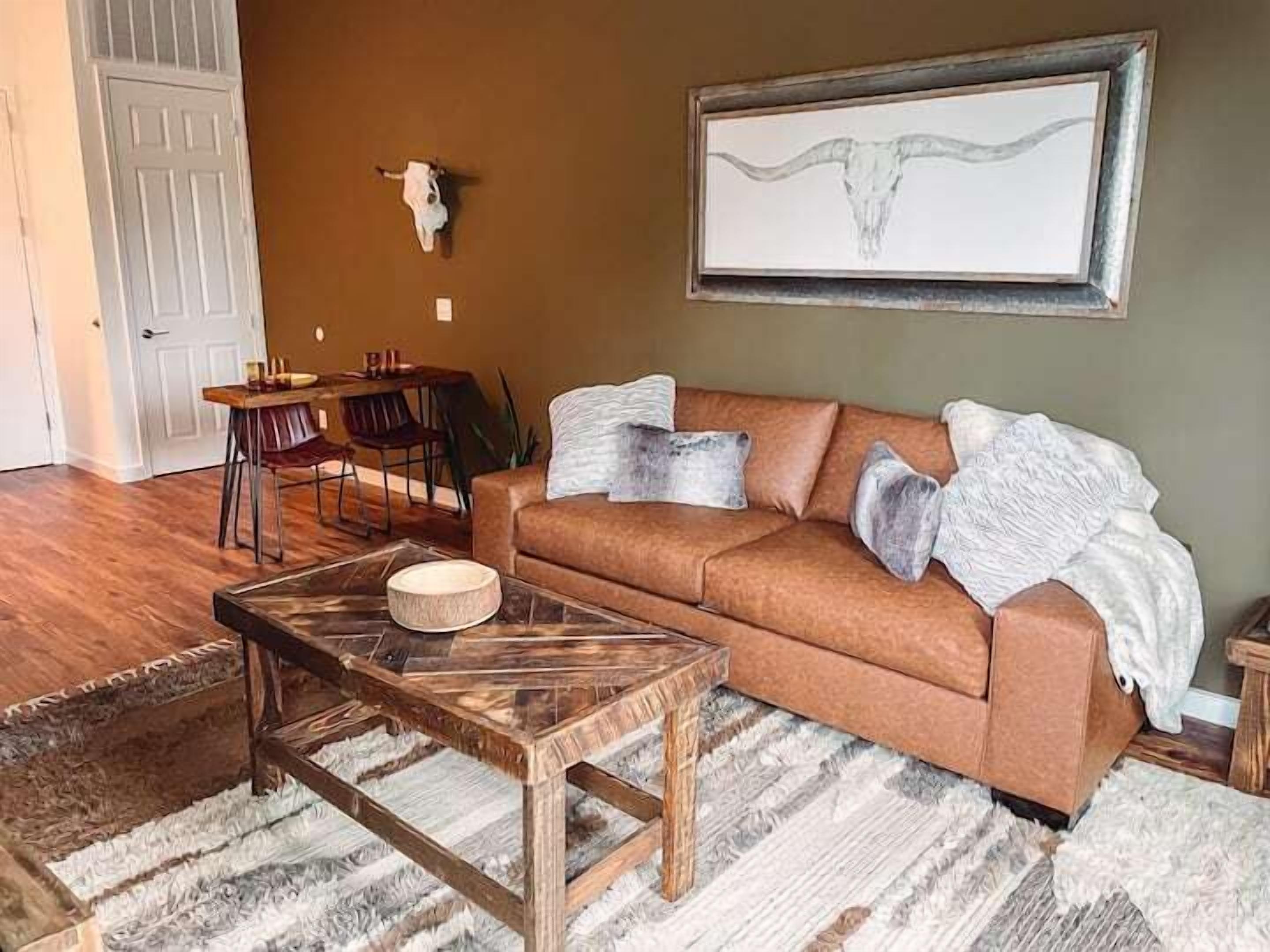 A living room with a brown sofa, a wooden coffee table, a rug, a dining area with chairs, and a wall-mounted artwork of a bull's head.