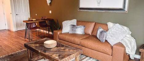 A living room with a brown sofa, a wooden coffee table, a rug, a dining area with chairs, and a wall-mounted artwork of a bull's head.