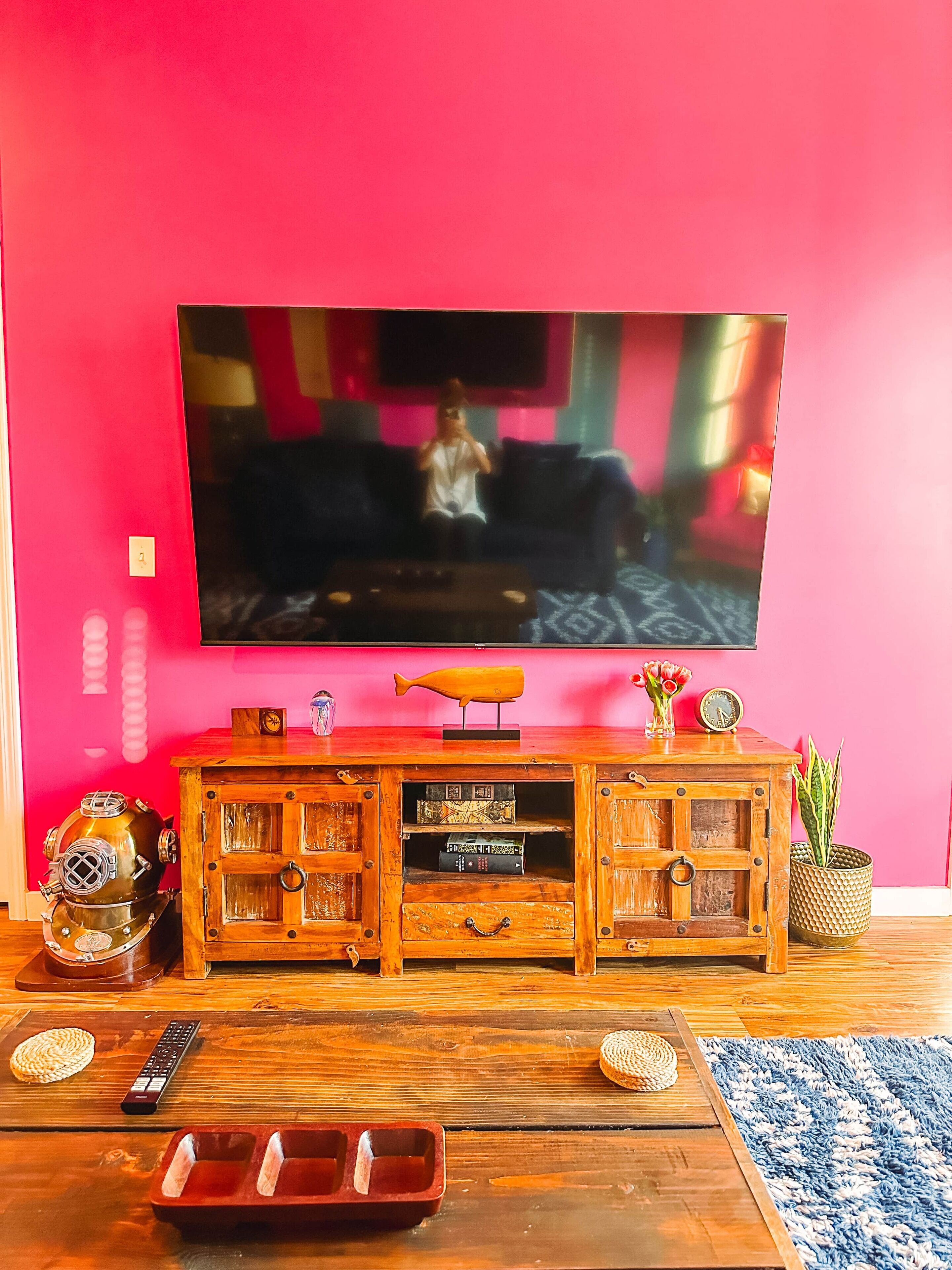 A living room with a TV mounted on a pink wall, wooden entertainment unit with books and decorative items, and a blue rug on the floor.