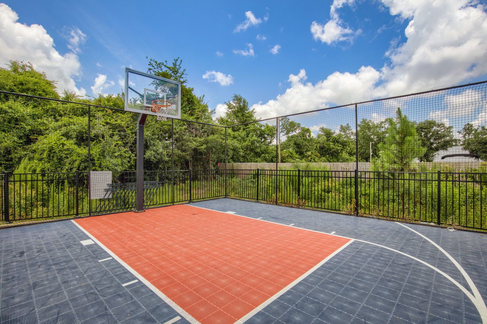 Een buitenbasketbalveld met een net, omgeven door een hek en bomen.