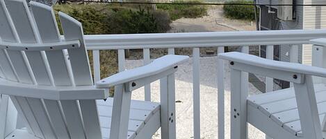 Un terrazzo in legno con ringhiere bianche che si affaccia su una spiaggia, con due poltrone Adirondack bianche e vista su una casa e l'oceano.