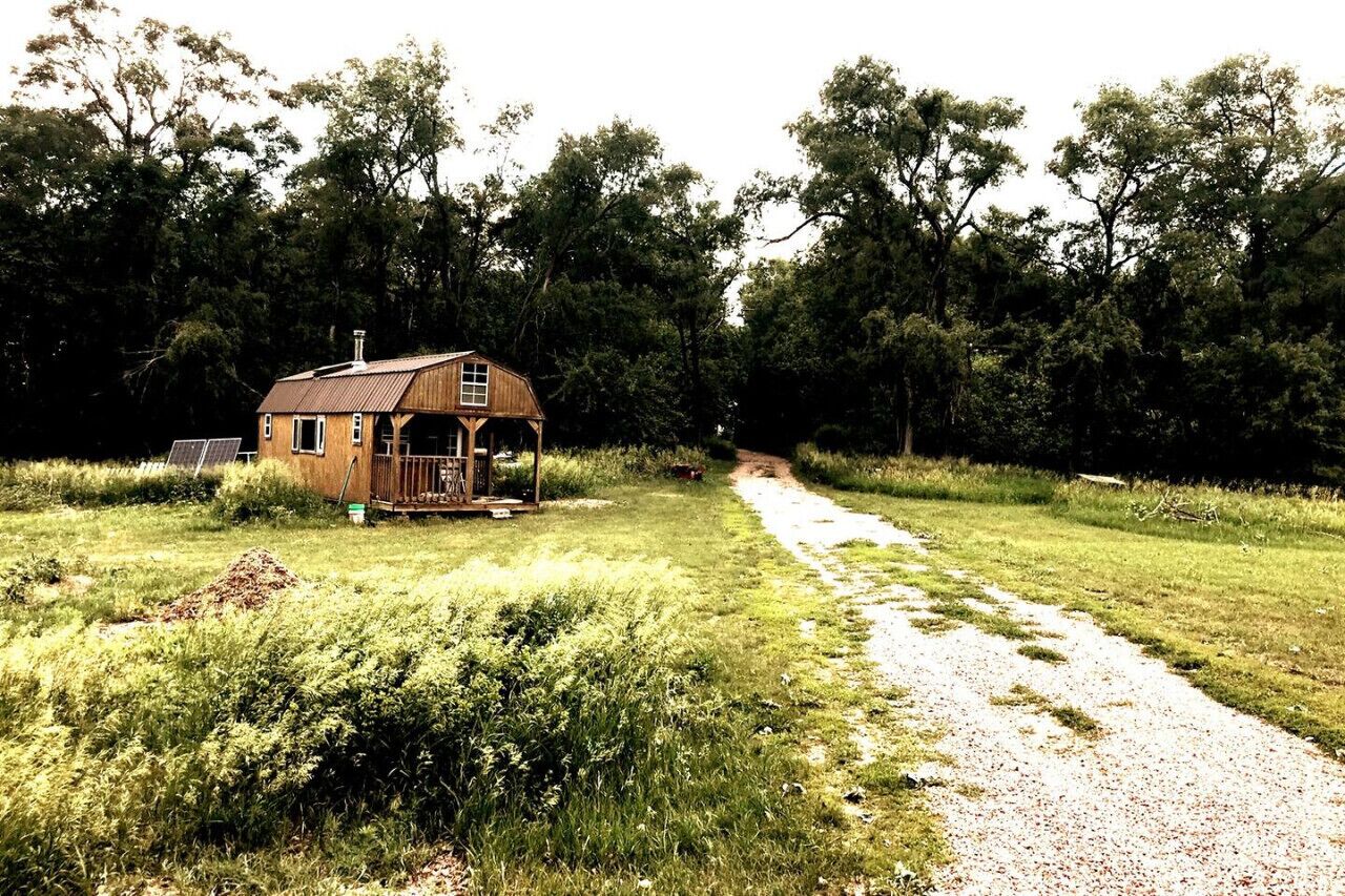 Une cabane en bois rustique, dotée d’un porche, entourée d’une forêt dense et accessible par un chemin de gravier.