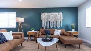 Un salon moderne avec un mur d’accent bleu foncé, une décoration murale en macramé et un canapé d’angle en cuir marron.