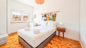 A modern bedroom with a large bed, wooden flooring, a window with a city view, a hanging light fixture, and a wall art piece. - GuestReady - Spacious Flat in the heart of Lisbon (Lisbon)