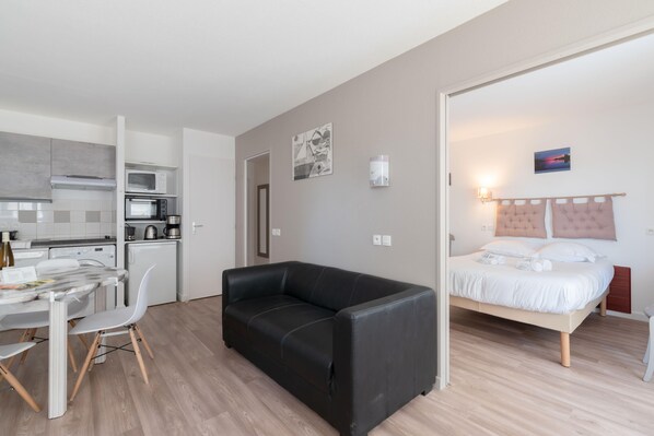 A modern apartment with a kitchenette, a black leather sofa, a dining area with a table and chairs, and a bedroom with a bed and a nightstand. - Le Green - Piscine - Mer - Wifi (Pornic)