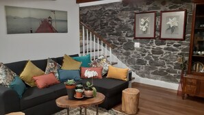 A living room with a black sofa, colorful cushions, a small round table with plants, a wooden stool, a staircase with a stone wall, and framed artwork on the wall. - Recanto das Florenças (3) – Stone House (Arco da Calheta)