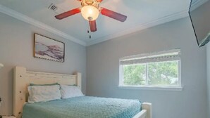 Un dormitorio con cama, mesita de noche, ventilador de techo, ventana y un cuadro en la pared.