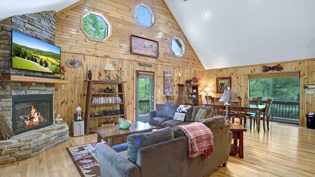A cozy living room with a stone fireplace, wooden walls, a large TV, and a dining area with a view of the outdoors. - Vacation at a family & pet friendly cabin in the Blue Ridge mountains! (Murphy)