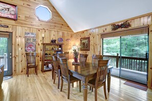 A wooden interior with a dining table, chairs, and a large window overlooking a forest.
