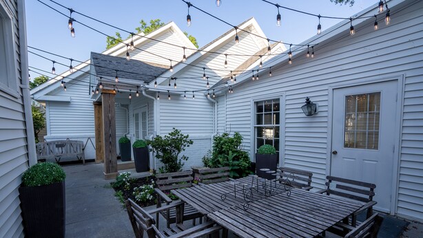 Une terrasse avec une table en bois et des bancs, entourée de plantes en pot et illuminée de guirlandes lumineuses.