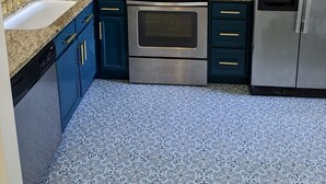 Une cuisine avec des meubles blancs, un four à micro-ondes, un four et un réfrigérateur en acier inoxydable, des meubles bleus, des comptoirs en granit et des carreaux de sol à motifs.