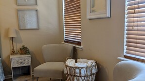 Une pièce chaleureuse avec un fauteuil de couleur claire, un panier en osier rempli de serviettes, une petite table d’appoint avec une lampe et des œuvres d’art encadrées accrochées aux murs.