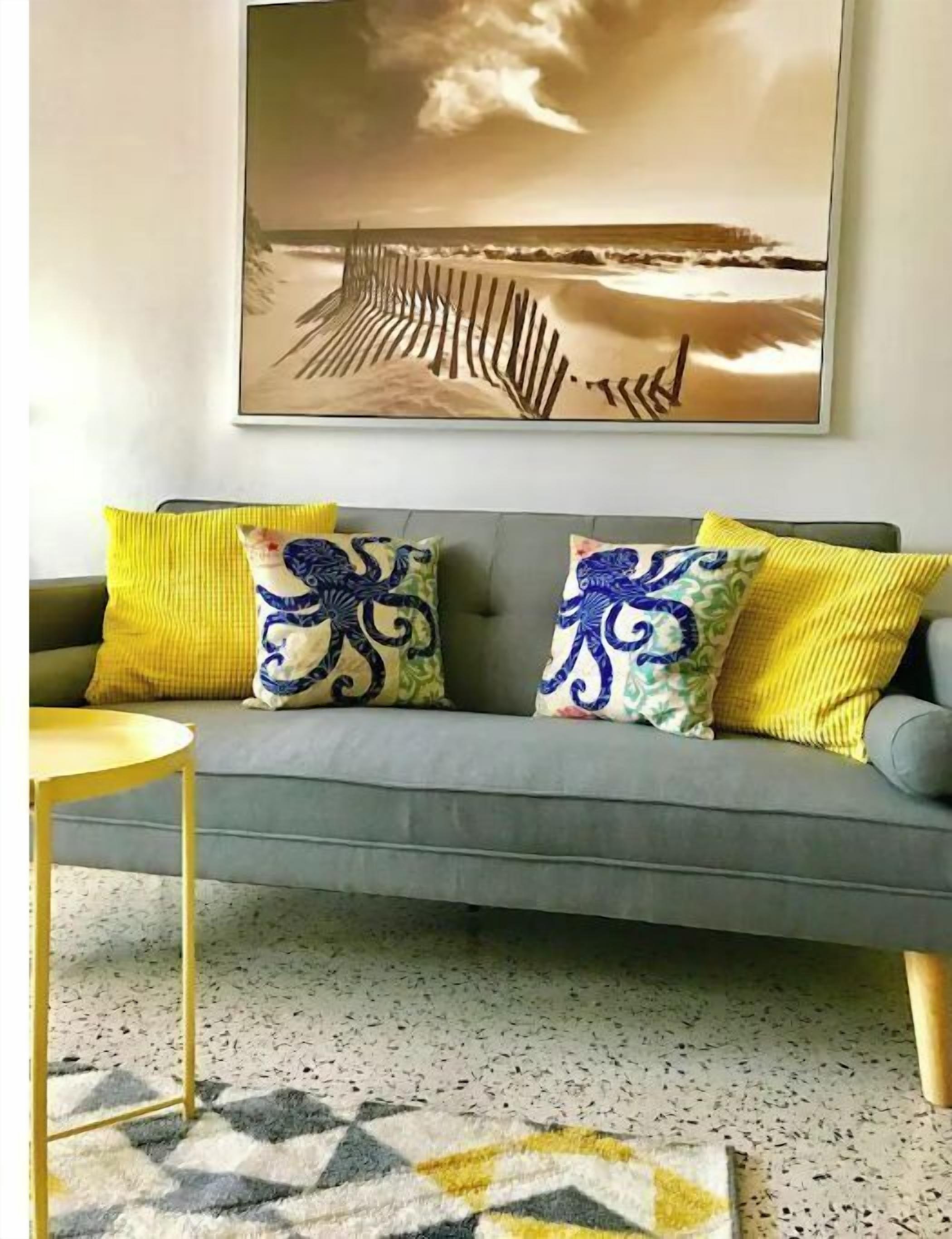 Un sofá gris con cojines estampados en amarillo y azul, una mesita redonda amarilla y una alfombra con diseño.