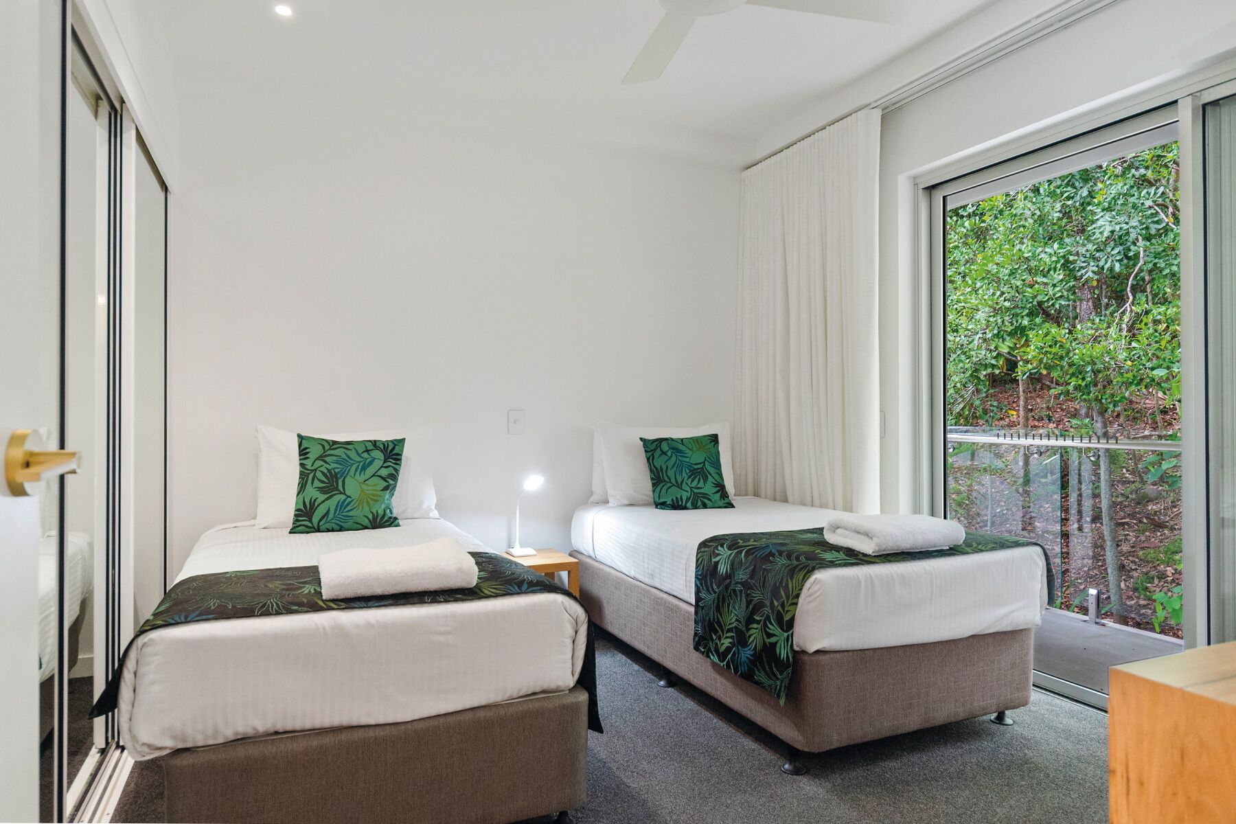 A hotel room with two single beds, each with a green leaf-patterned pillow and a white towel. The room has a large window with a view of greenery outside.