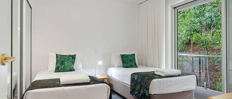 A hotel room with two single beds, each with a green leaf-patterned pillow and a white towel. The room has a large window with a view of greenery outside.