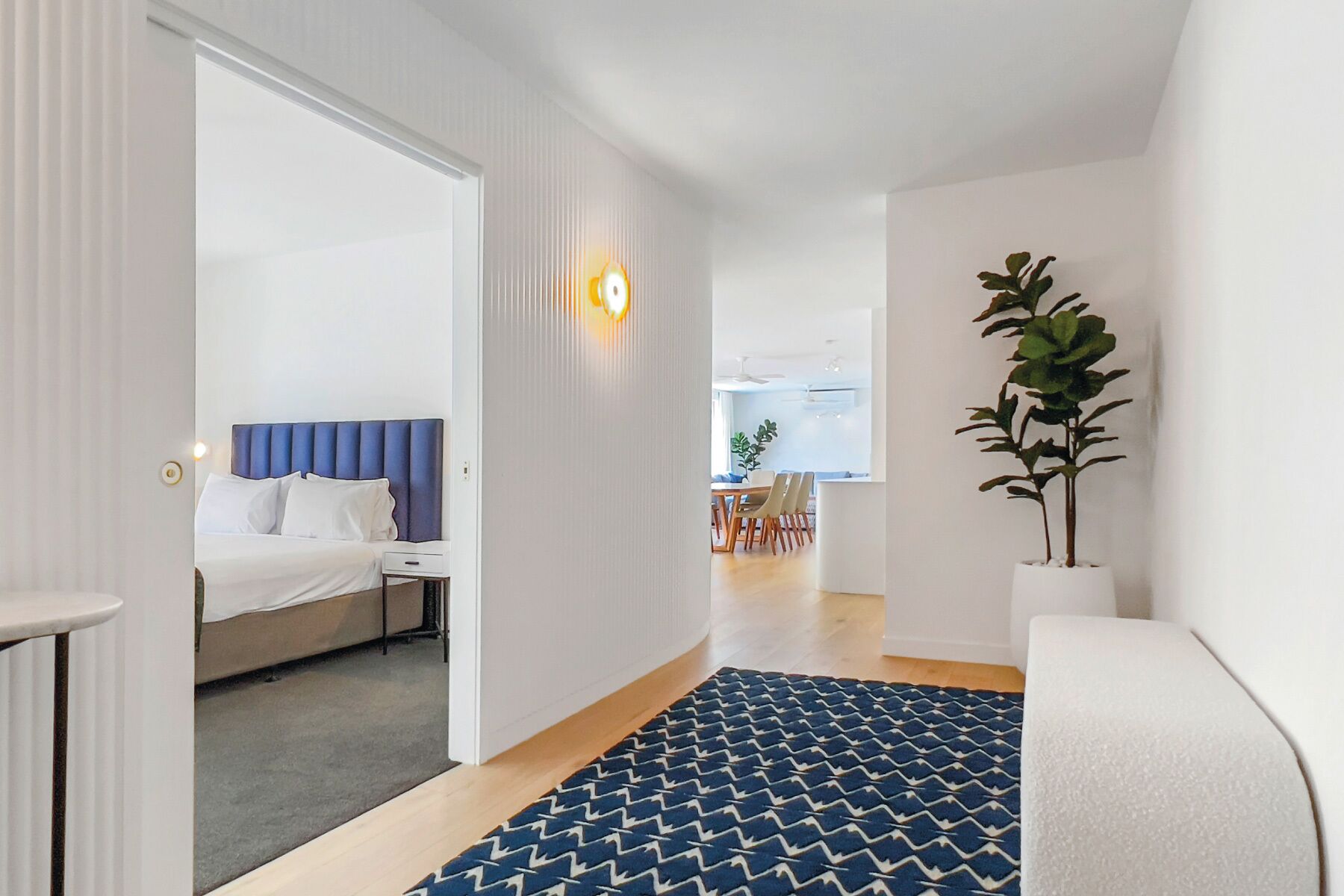 Une chambre d’hôtel avec un lit, un bureau et une chaise. Il y a une plante dans un coin et une fenêtre avec des rideaux.
