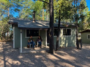 A small cabin with a porch, two people sitting, and a tree in front. - Cabin 8 — Romantic Queen Cabin with Private Indoor Hot Tub (Lakeside)