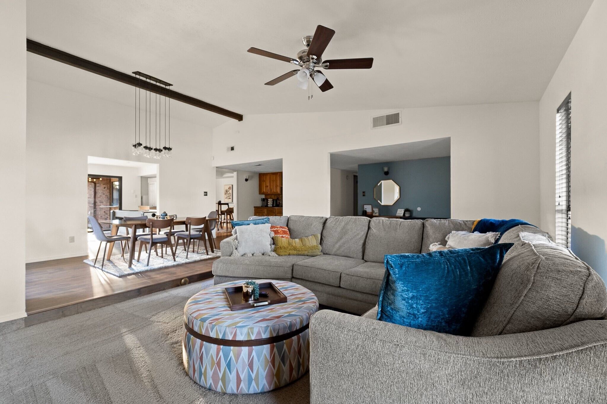 A spacious living room with a large gray sofa, a patterned ottoman, and a ceiling fan. There is a dining area in the background with a wooden table and chairs.