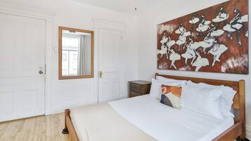 A bedroom with a large bed, white walls, a wooden headboard, a nightstand, a mirror, and a painting.