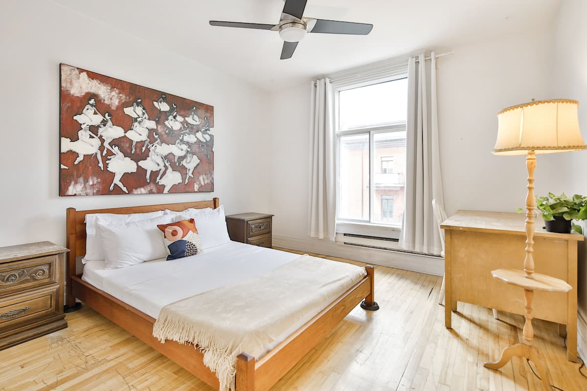 A neatly arranged bedroom with a wooden bed, a bedside table with a lamp, a nightstand, and a large window with curtains.