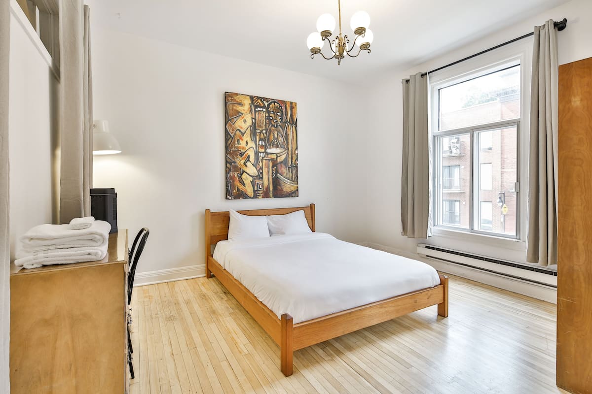 A bedroom with a wooden bed, a painting on the wall, a window with curtains, and a chandelier.