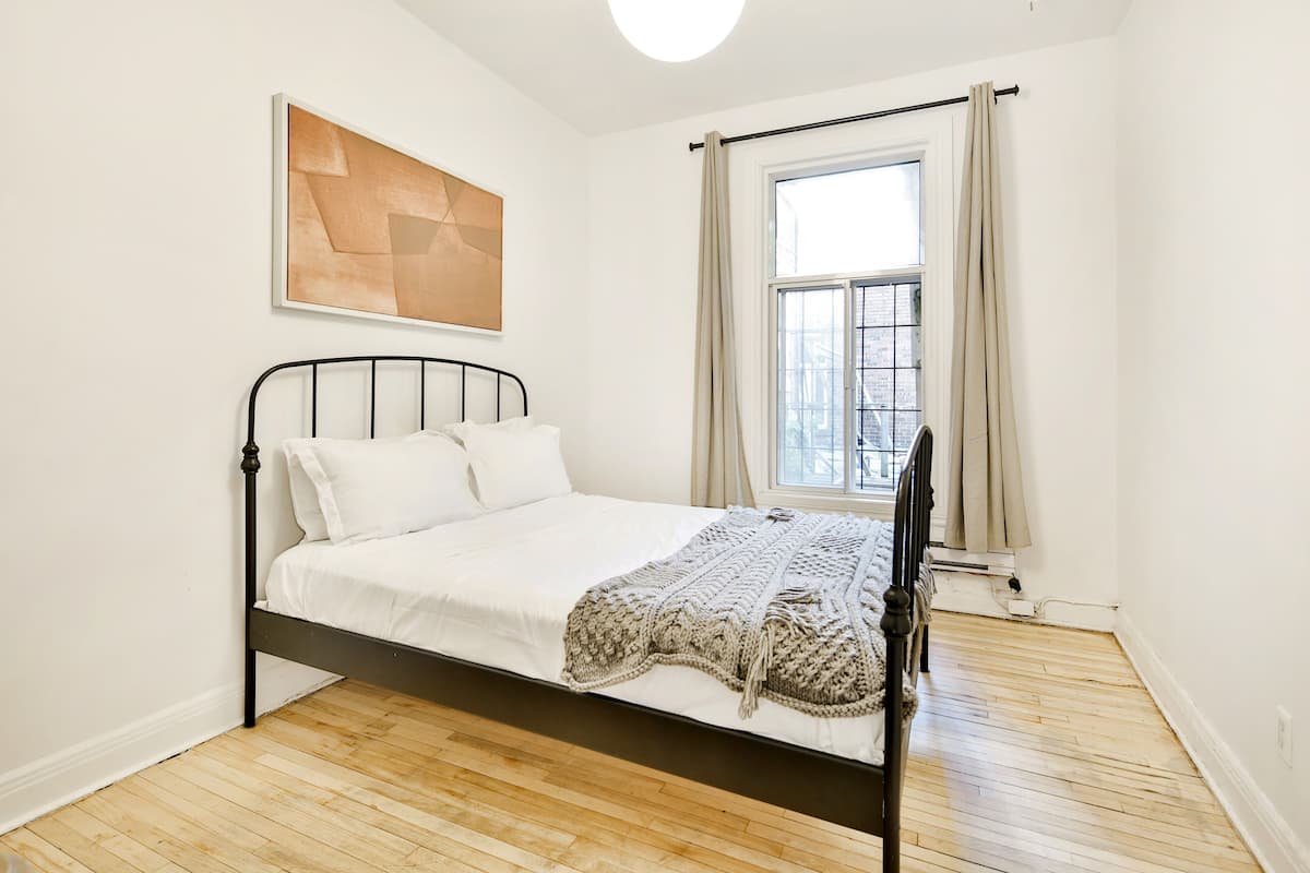 A bedroom with a bed, a window with curtains, a wooden floor, and a wall-mounted abstract painting.