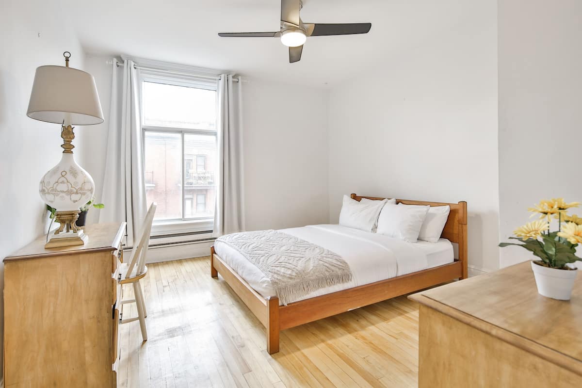 A bedroom with a wooden bed, a desk with a lamp, a ceiling fan, and a window with curtains.