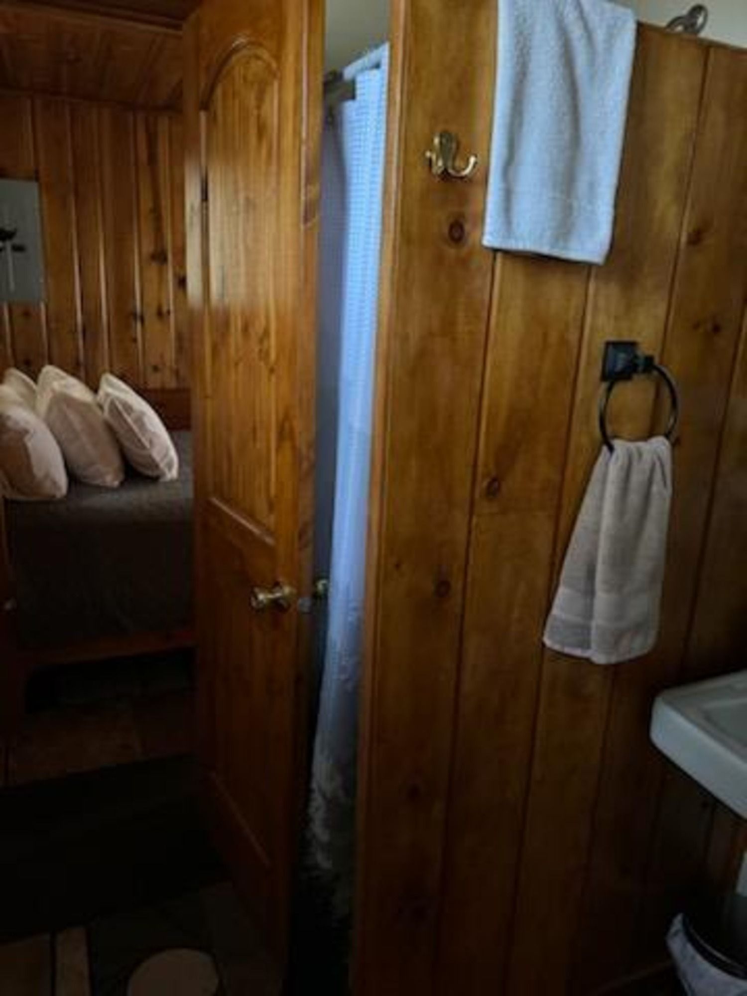 Ein Zimmer mit Holzboden, einem Bett, einer Tür und einem Badezimmer mit Waschbecken und Handtuchhalter.