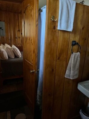 Um quarto de madeira com uma cama, porta e banheiro, incluindo pia e porta-toalhas.