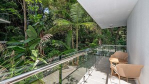 A balcony with glass railing, surrounded by lush greenery and two wicker chairs.