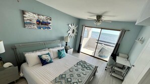 Une chambre à coucher comprenant un lit, une table de chevet, un bureau, une chaise, un ventilateur de plafond, une fenêtre donnant sur l’océan et un tableau accroché au mur.